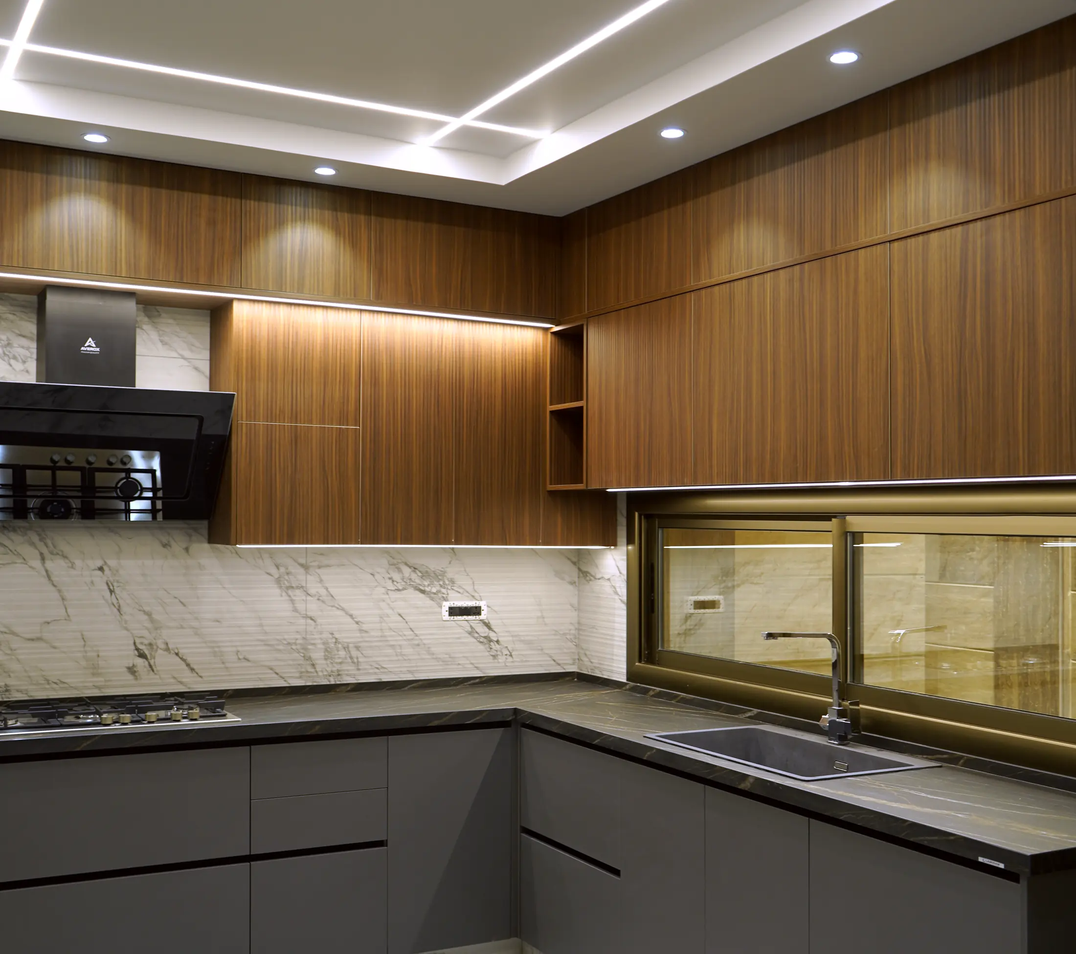 مطبخ من تنفيذ Wood Stone على شكل حرف L: خزائن علوية بخشب الجوز، ووحدات سفلية رمادية داكنة، وسطح عمل داكن بعروق، وإطارات إضاءة خطية في السقف